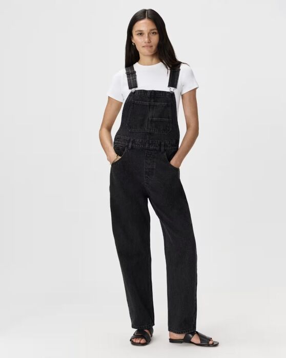 A person wearing a white t-shirt under black denim overalls and black slide sandals.