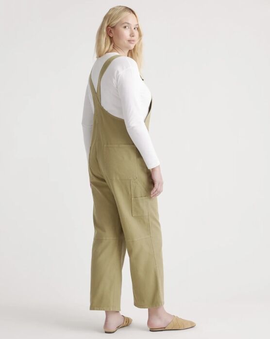 A woman wears a long-sleeved white shirt under olive green overalls with side pockets, paired with tan mules.
