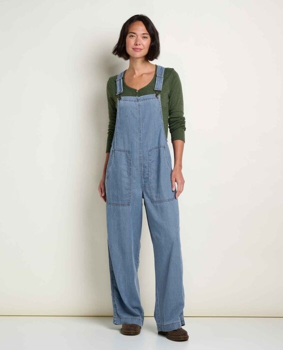 A woman wearing light-wash denim overalls over a dark green long-sleeved shirt.
