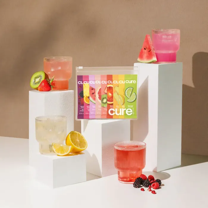 An arrangement of colorful drink packets and glasses filled with various fruit-infused beverages on geometric white blocks, surrounded by sliced kiwi, lemon, and berries.