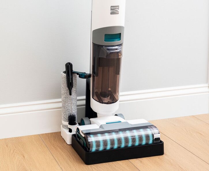 Cordless vacuum cleaner with a water-filled tank and cleaning brush attachment on a wooden floor against a wall.