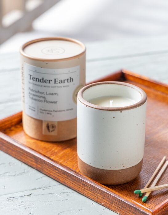 A white and brown ceramic candle sits on a wooden tray with a labeled candle container and some matches nearby.