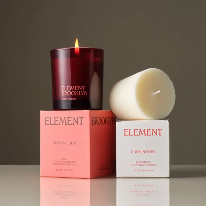 A lit burgundy candle in a glass holder sits on a pink box next to a white candle refill on a white box, both labeled “Element Brooklyn Dusk in Eden.”.