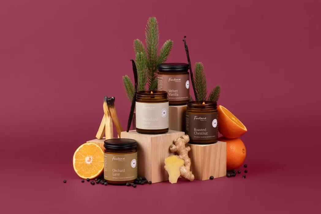 Four candles in glass jars displayed with wooden blocks, oranges, ginger, pine branches, and vanilla beans against a maroon background.