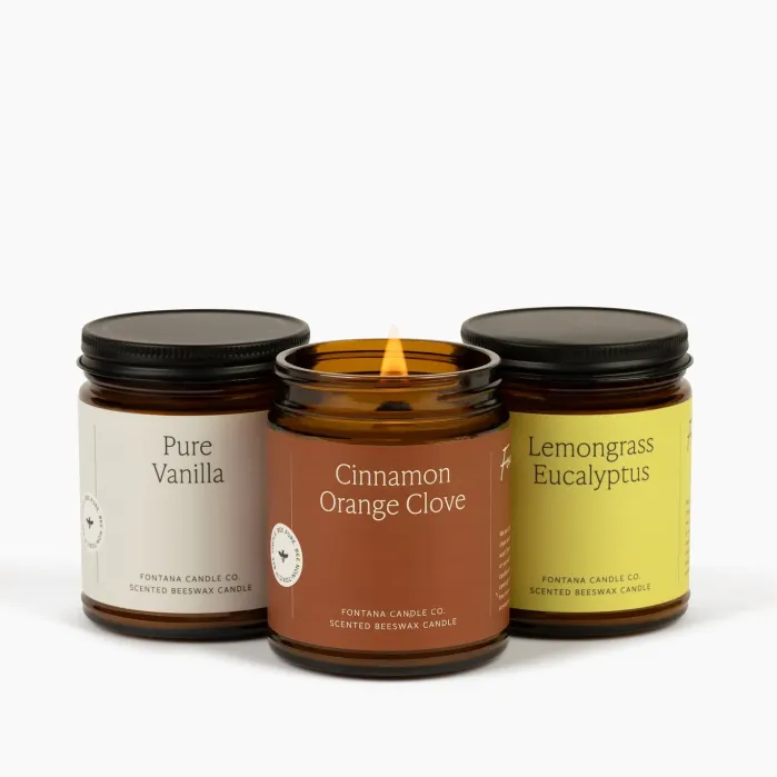 Three scented candles in glass jars labeled Pure Vanilla, Cinnamon Orange Clove (lit), and Lemongrass Eucalyptus are arranged side by side against a white background.