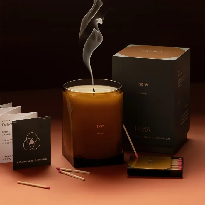 A brown glass candle labeled “hara (home)” sits lit with smoke above the wick, next to an open matchbox, burnt matches, instruction cards, and the candle’s packaging box.