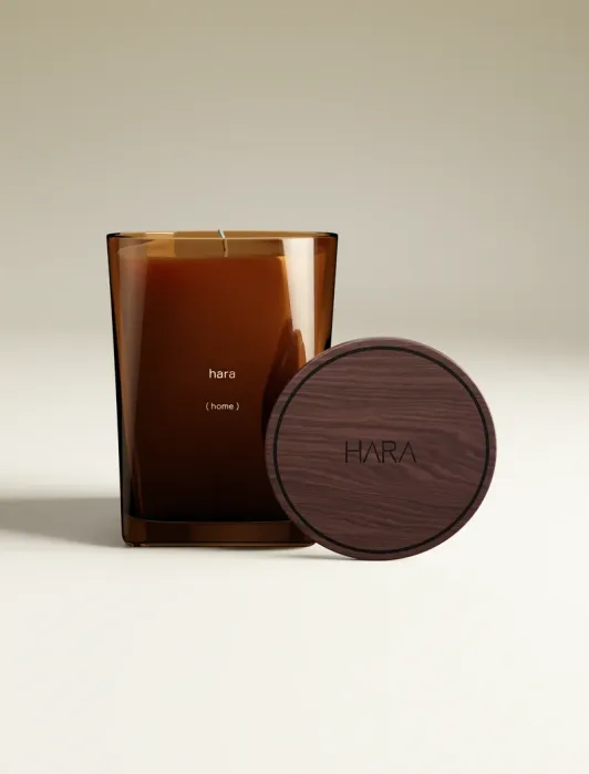 A brown glass candle labeled “hara (home)” sits on a white surface, with its wooden lid marked “HARA” leaning against it.