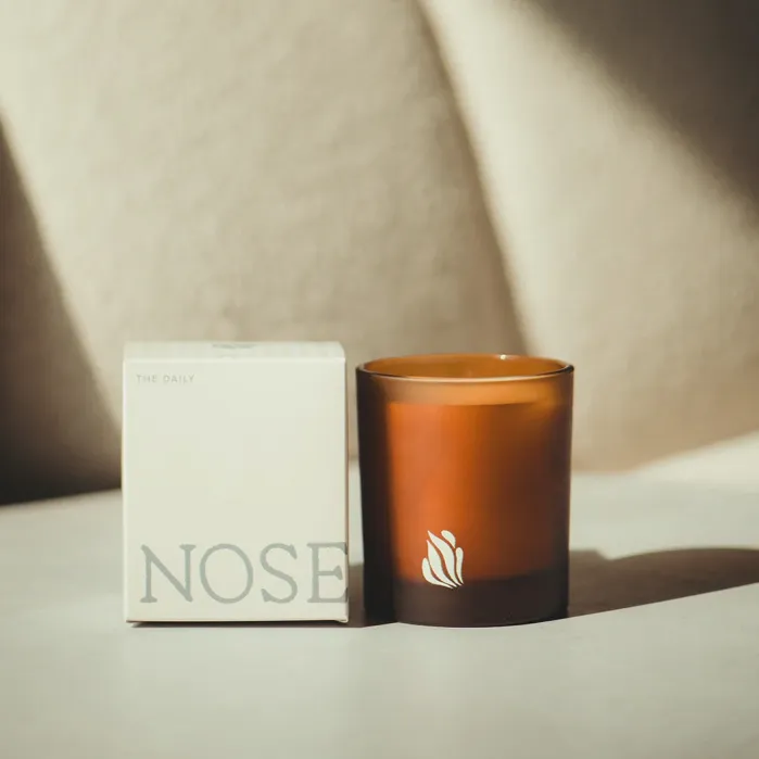 A brown glass candle with a white logo sits next to a box labeled “NOSE” in large gray letters, both on a light surface with soft sunlight and shadows.
