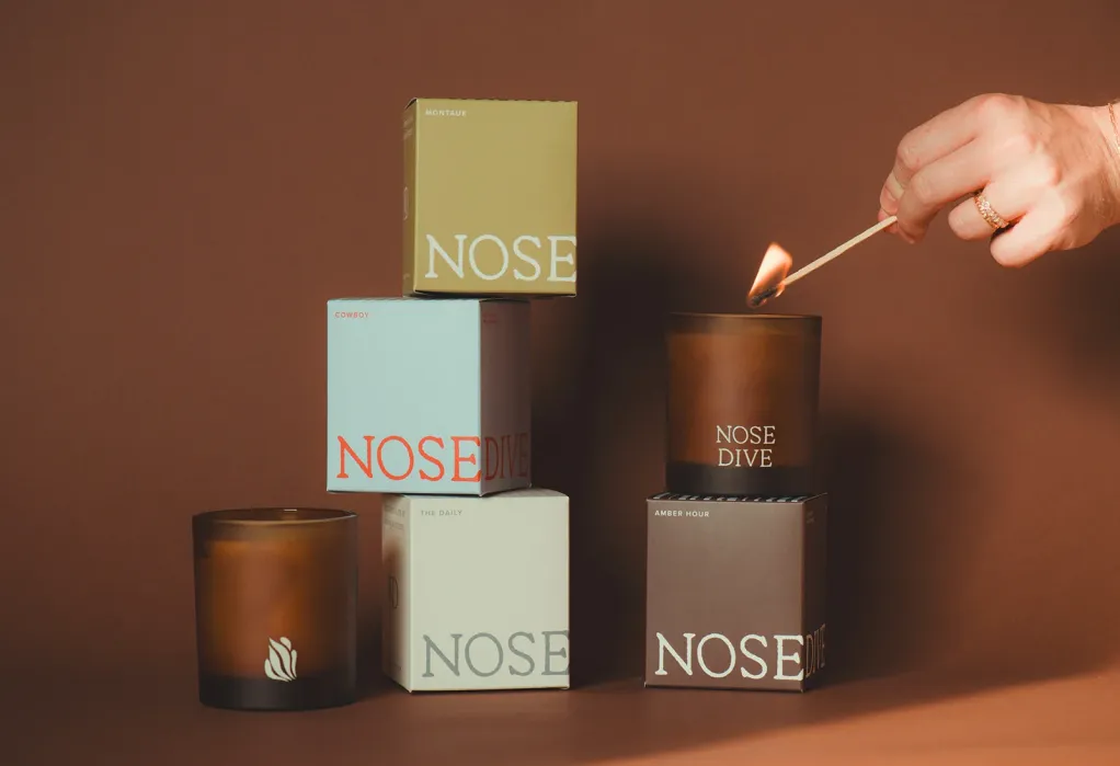 A hand lights a brown candle with a match; four boxed candles labeled “NOSE DIVE” are stacked beside it. The background is brown.