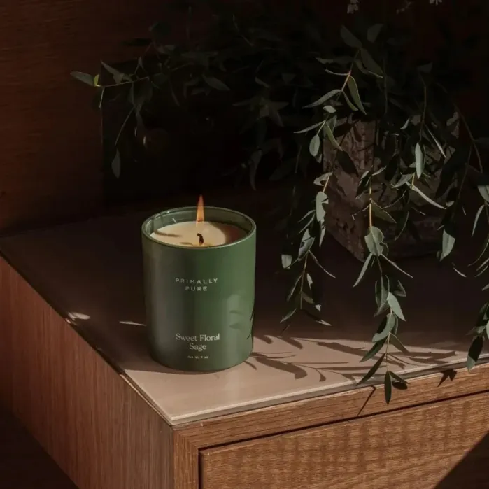 A green “Sweet Floral Sage” candle is lit on a wooden surface next to some leafy branches, with soft sunlight casting shadows.