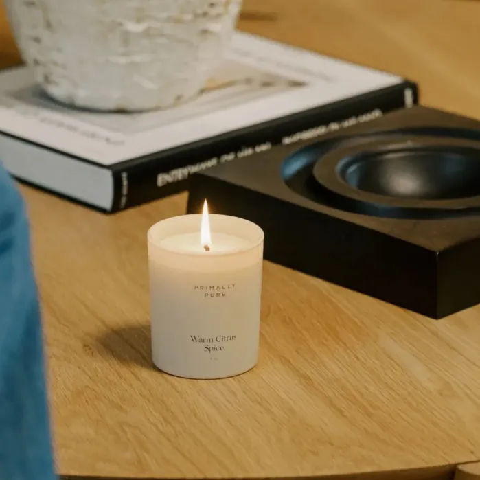 A lit candle labeled “Warm Citrus Spice” sits on a wooden table next to a black dish, a book, and a white textured pot.