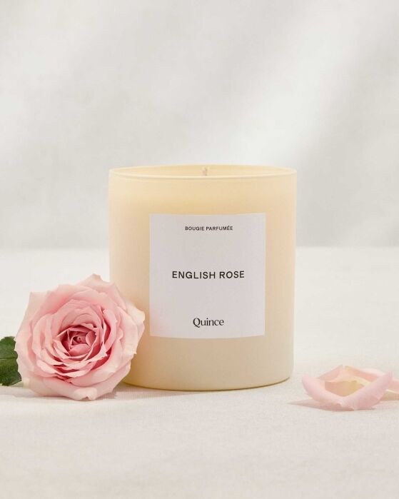 A cream-colored candle labeled “English Rose” sits beside a pink rose and its petals on a white surface with a soft background.