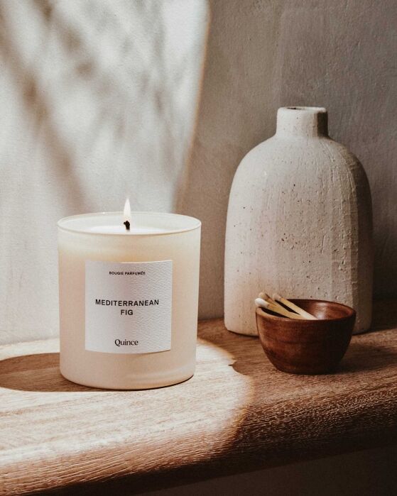 A lit Mediterranean Fig candle sits on a wooden surface next to a small bowl of matches and a textured white vase against a light wall.
