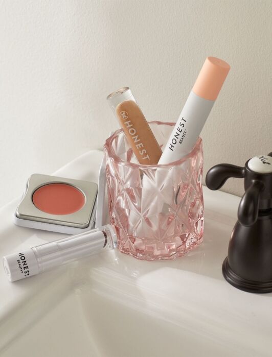 Cosmetic products including lip gloss, concealer, and a compact blush placed on a bathroom sink next to a pink glass.
