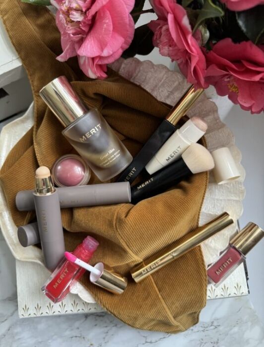 Makeup products, including a foundation bottle, blush, brushes, lipstick, and lip gloss, are arranged on a brown fabric with pink flowers nearby.