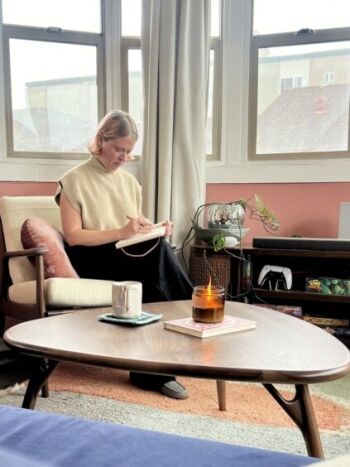 Person sitting on a chair, writing in a notebook. A mug and lit candle are on the table in front. Large windows in the background.