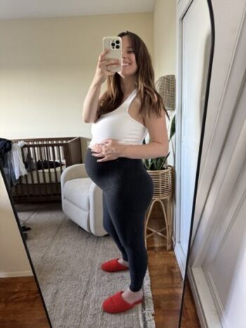 A woman takes a mirror selfie in a bedroom, wearing a white top, black leggings, and red slippers, holding her pregnant belly. A crib and chair are visible in the background.