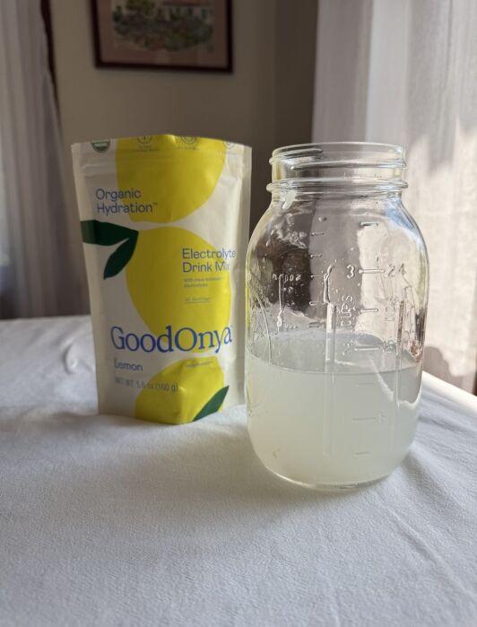 A glass jar with a cloudy lemon drink sits in front of a GoodOnYa Lemon Organic Hydration Electrolyte Drink Mix packet on a white table.