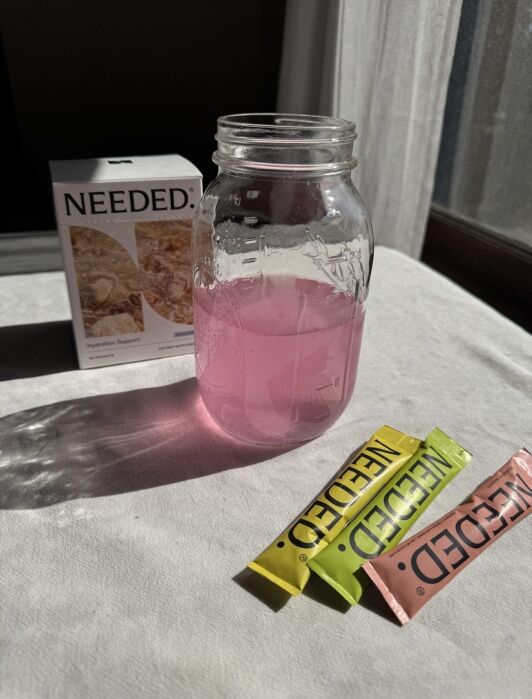 A glass jar with pink liquid sits on a table beside three colorful Needed. supplement packets and a Needed. Hydration Support box. Sunlight streams in from a nearby window.