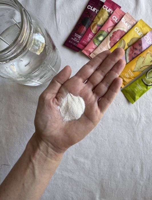A hand holding a small mound of white powder, with a glass jar of water and several colorful electrolyte drink mix packets in the background.