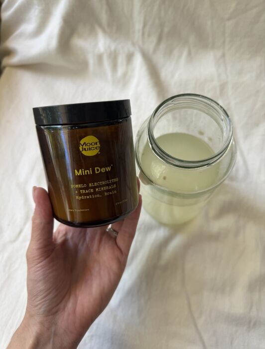 A hand holds a jar labeled "Moon Juice Mini Dew," next to a glass jar containing a light yellow liquid, both set against a light-colored fabric background.