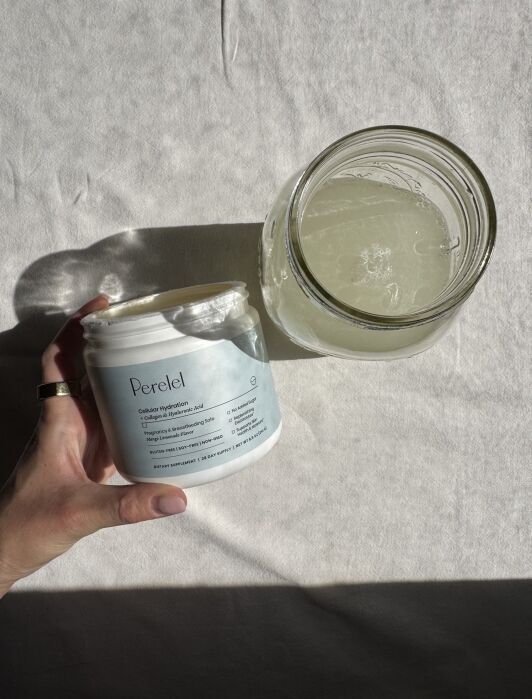 A hand holds a container of Perelel Cellular Hydration powder next to a glass jar filled with a pale liquid on a light-colored fabric surface.