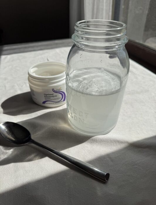 A spoon, an open container of moisturizer, and a glass mason jar filled with cloudy water are arranged on a white tablecloth in natural light.