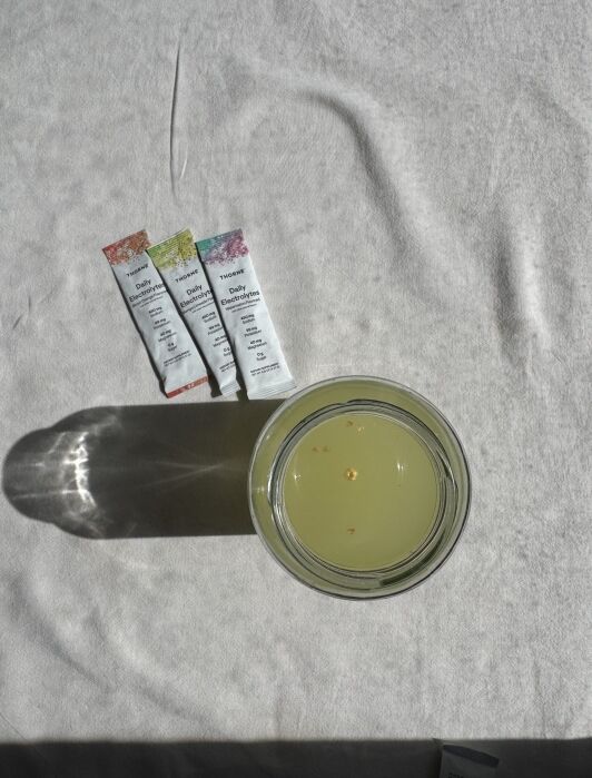 A glass of yellow liquid sits on a white surface next to three packets labeled “Daily Electrolytes.” Shadows are cast from the glass and packets.