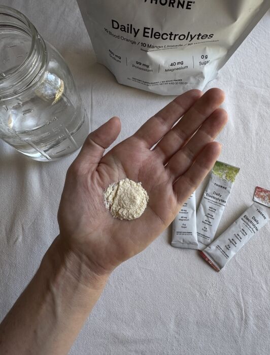 A hand holds a small pile of powdered electrolyte supplement; nearby are a glass of water, electrolyte packets, and a large bag labeled "Daily Electrolytes.