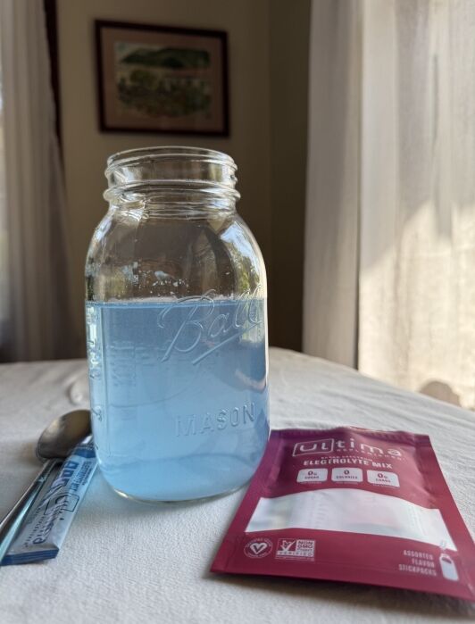 A mason jar filled with blue liquid sits on a table beside a spoon, an empty stir stick wrapper, and an Ultima electrolyte mix packet.