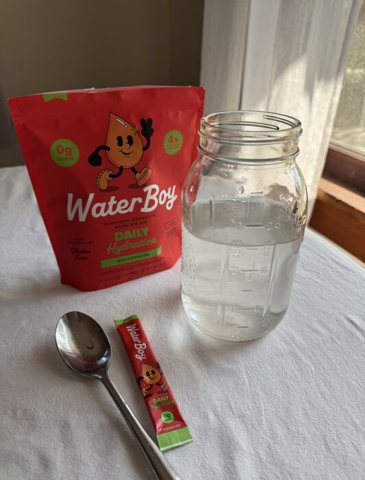 A packet of WaterBoy hydration powder, an empty packet, a spoon, and a mason jar filled with water are arranged on a white surface near a window.