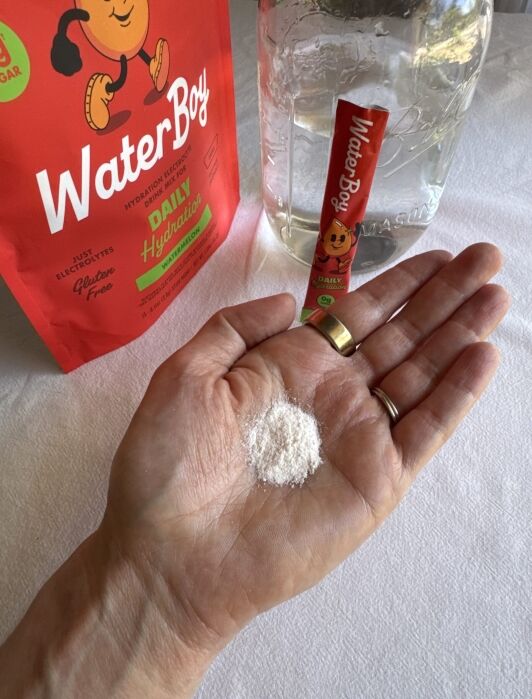 A hand holds a small pile of white powder. Nearby are a red WaterBoy hydration packet, a red WaterBoy bag, and a glass jar of water on a white tablecloth.
