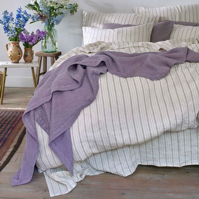 A bed with striped bedding and pillows, a textured purple blanket draped over it, and vases with flowers on a small table beside the bed.