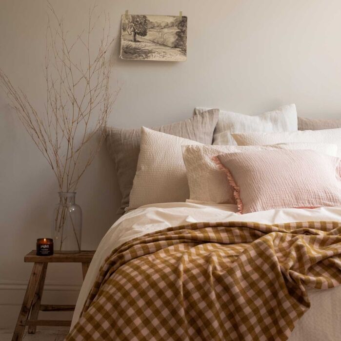 A neatly made bed with neutral-toned pillows and a brown gingham blanket, a wooden stool with a vase and candle, and a small artwork hanging on a pale wall.