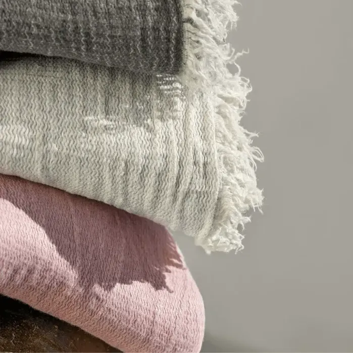 A close-up of three folded textured blankets stacked on top of each other in gray, off-white, and pink colors. The off-white blanket has a fringed edge.
