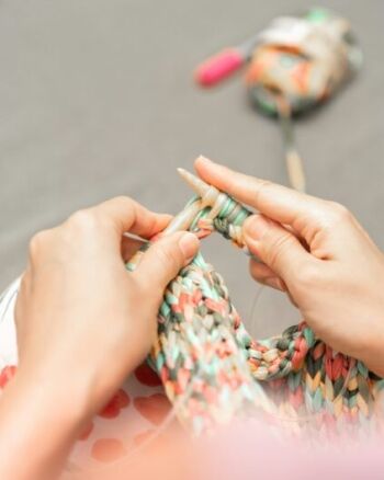 A person knitting with multicolored yarn, using wooden needles, with a ball of yarn in the background.