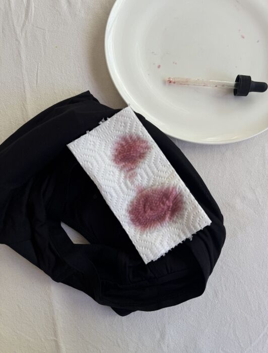 A fabric with two reddish stains on a white paper towel, next to a dropper and an empty white plate, placed on a white surface.
