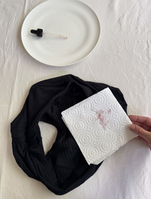 Black fabric with a small red stain next to a white paper towel and a dropper on a white plate, all placed on a white surface.