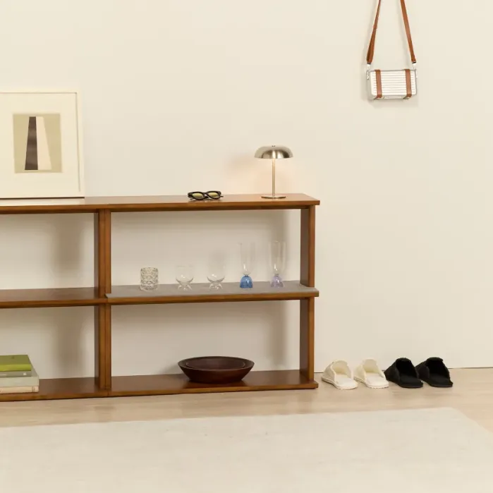 A wooden shelf with books, glasses, a lamp, and decor items stands against a beige wall; two pairs of slippers and a small bag hang nearby.
