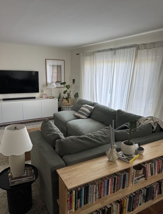 A living room with a green sectional sofa, a large TV on a white cabinet, sheer curtains, houseplants, and a bookshelf filled with books behind the sofa.