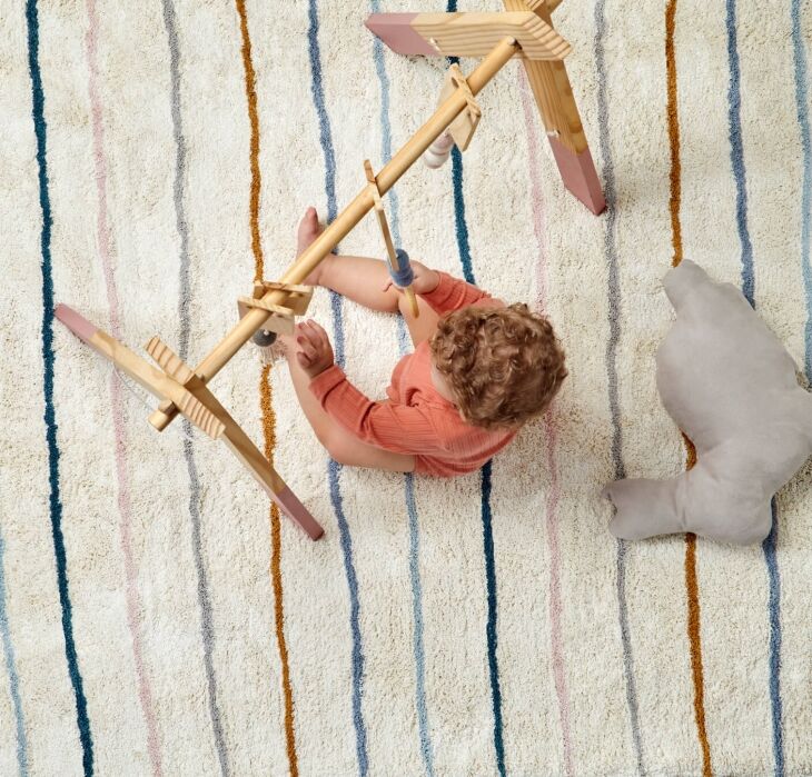 A baby sits on a striped rug, playing with a wooden play gym and surrounded by a plush toy.