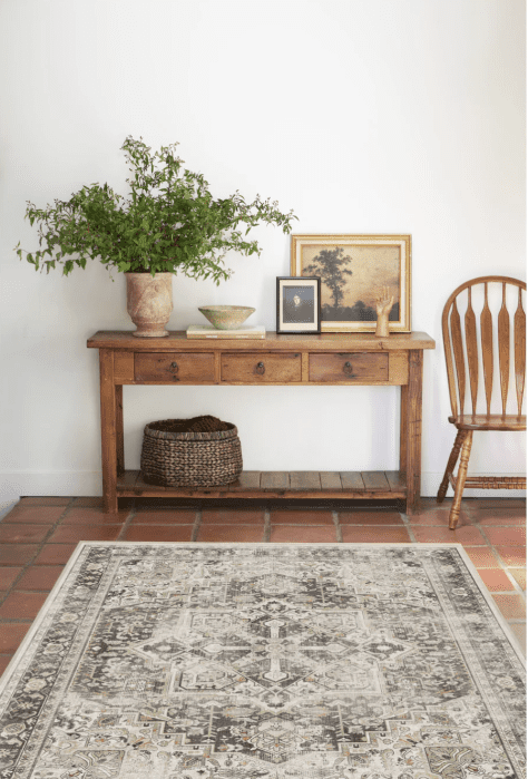 Wooden console table with plants, framed pictures, and decorative items. A wicker basket is underneath. A patterned rug lies on terracotta tiles next to a wooden chair.