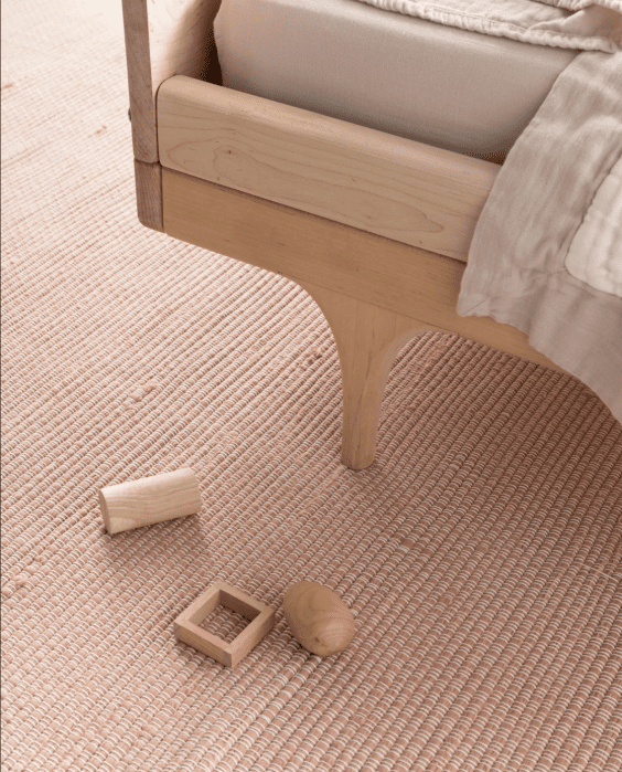 Wooden bed frame on a textured pink rug with three wooden toys: a cylinder, a cube with a square hole, and an egg-shaped piece nearby.