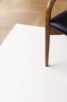 The corner of a wooden chair with a black seat is on a light-colored rug on a wooden floor.