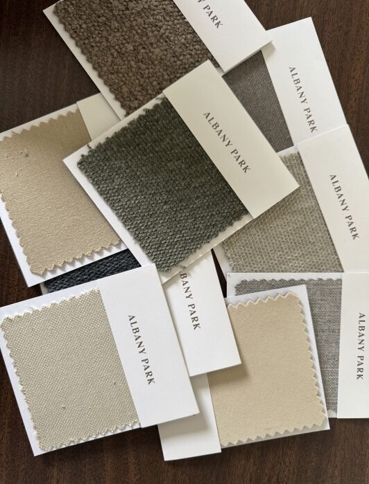 A scattered arrangement of Albany Park fabric swatches in various neutral tones, including beige, gray, and taupe, displayed on a dark wood surface.