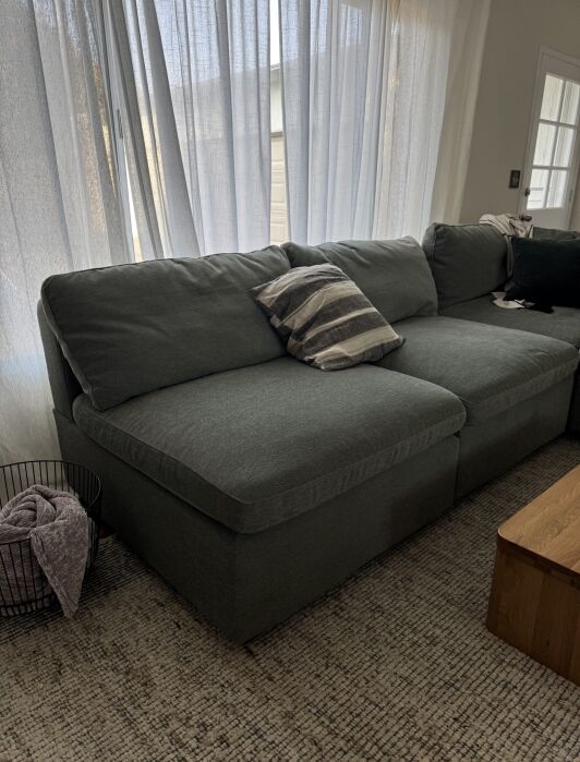 A green sectional sofa with two cushions and a striped pillow sits in a living room with light curtains, a rug, a wooden table, and a black wire basket with a blanket.
