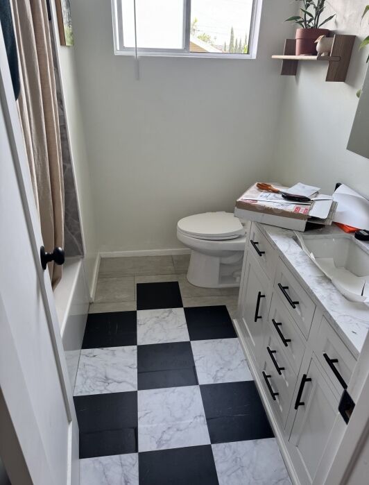 A bathroom with a black and white checkered floor, white toilet, marble countertop with drawers, and a window above the toilet. Some items are placed on the counter.