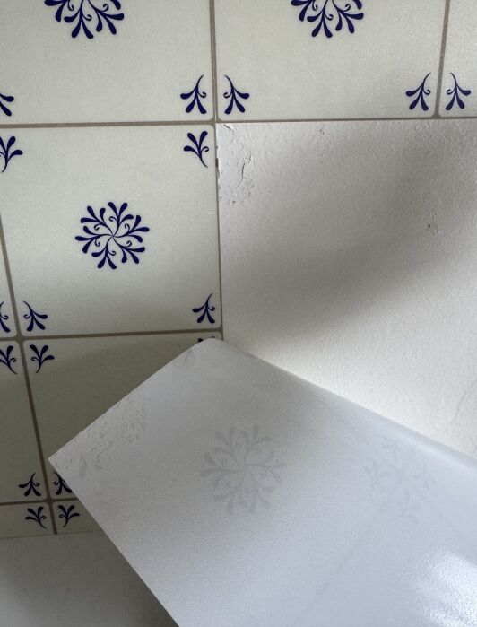 A decorative tile sticker is partially peeled back from a tiled wall, revealing a plain, damaged white surface beneath patterned tiles.