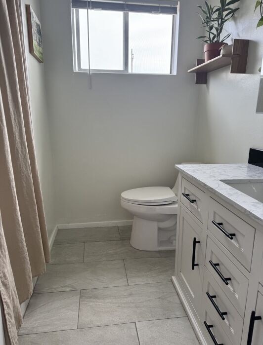 A small bathroom with a toilet, white vanity with drawers, tiled floor, window with blinds, and a plant on a wall shelf.