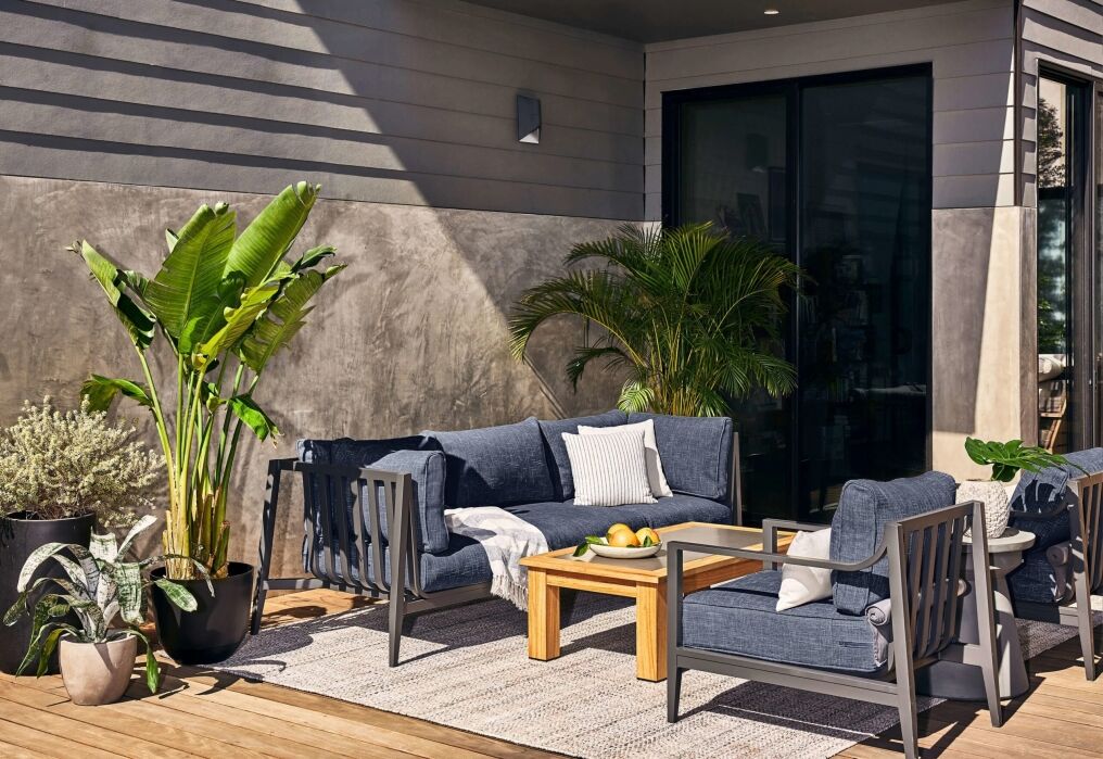 A modern outdoor patio with gray cushioned seating, a wooden coffee table, a rug, and several potted green plants against a house wall.